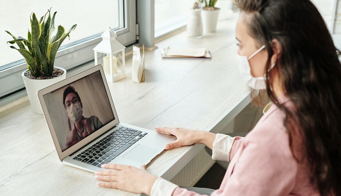 woman in pink shirt wearing mask video chatting through laptop with a man who is also wearing a surgical mask